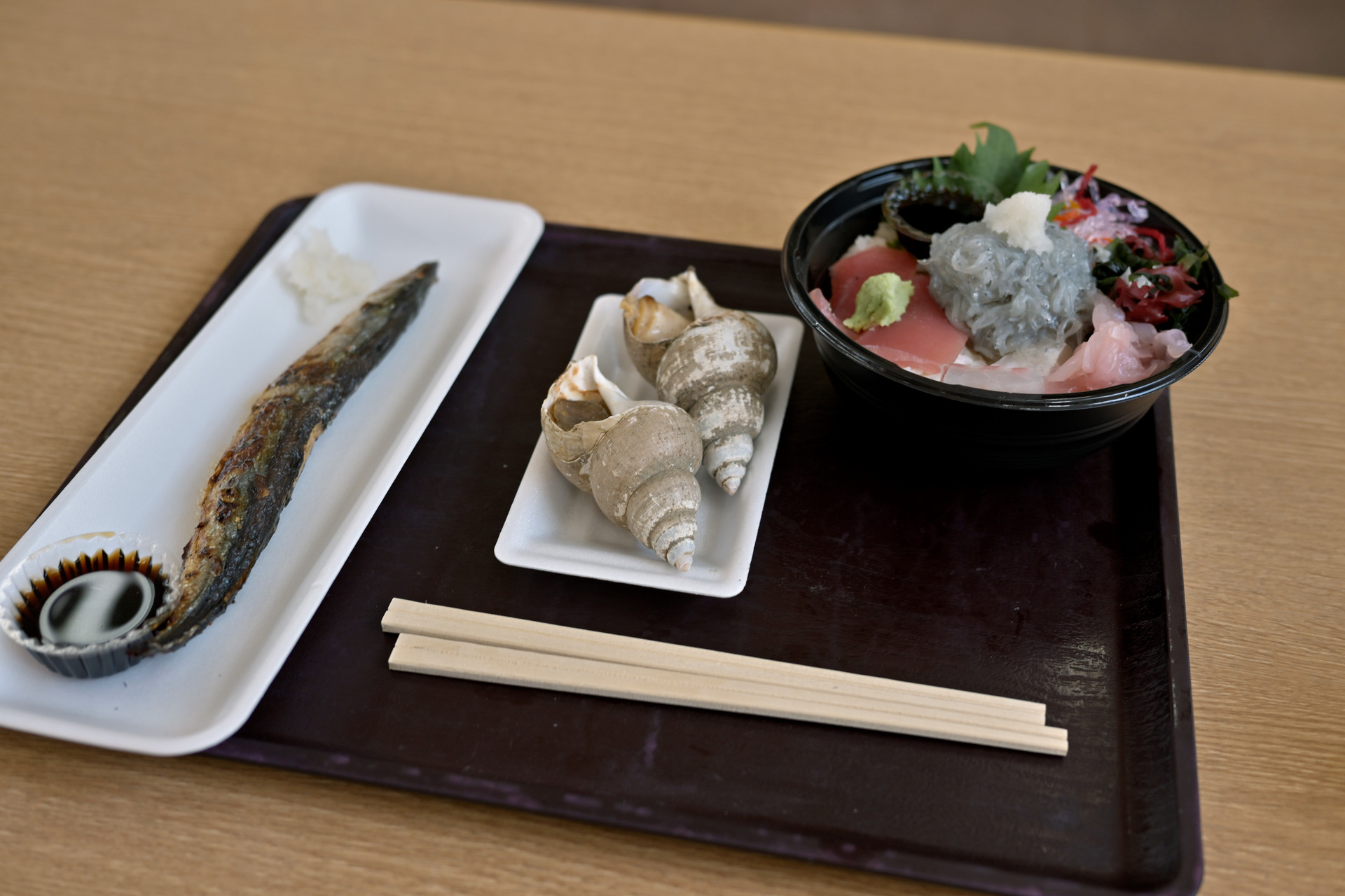 お盆の左にサンマの塩焼き、中央に殻付きのバイ貝2個、右に生しらすと刺身の載った丼が並ぶ