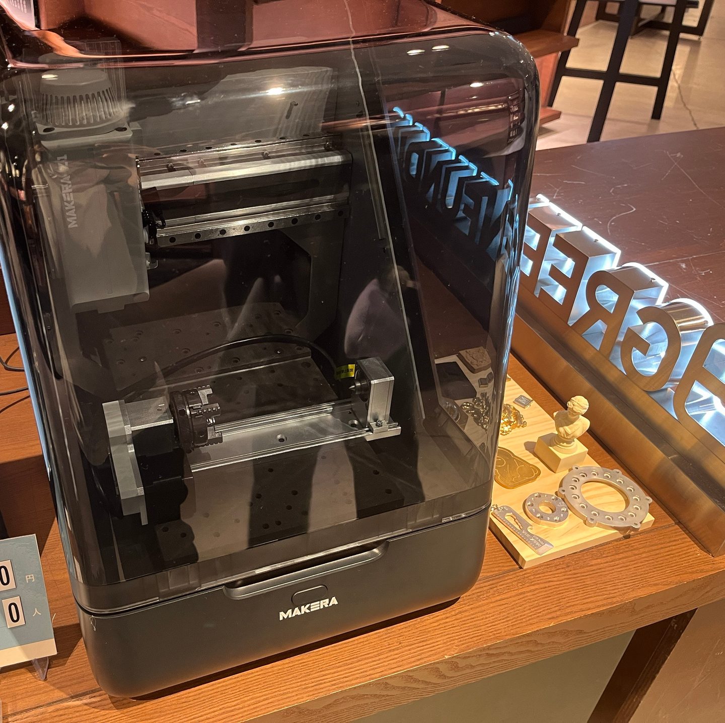 透明なカバーの中に4軸モジュールを取り付けたMakera Z1。横に、アルミや真鍮を削った見本の部品が並んでいる